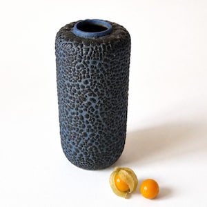 Op de afbeelding: Een cilindervormige keramische vaas met een getextureerde, donkerblauwe buitenkant en een gladde, lichtblauwe binnenkant. Twee kleine, oranje physalisvruchten liggen naast de vaas op een witte ondergrond. De vaas heeft een unieke, organische uitstraling.
