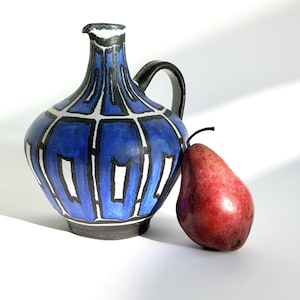 Peut inclure: Une cruche décorative bleue et noire avec une anse noire et un bord blanc et noir. Une poire rouge repose à côté de la cruche. La cruche présente un motif géométrique avec des formes rectangulaires blanches.