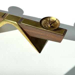K&ouml;nnte beinhalten: Ein Kerzenhalter im Mid-Century-Modern-Stil mit Messing- und Holzdesign. Der Kerzenhalter verf&uuml;gt &uuml;ber eine kleine, runde Messingmulde f&uuml;r die Kerze und einen geometrischen Holzsockel. Das Holz ist dunkelbraun und das Messing hat einen warmen, goldenen Farbton.