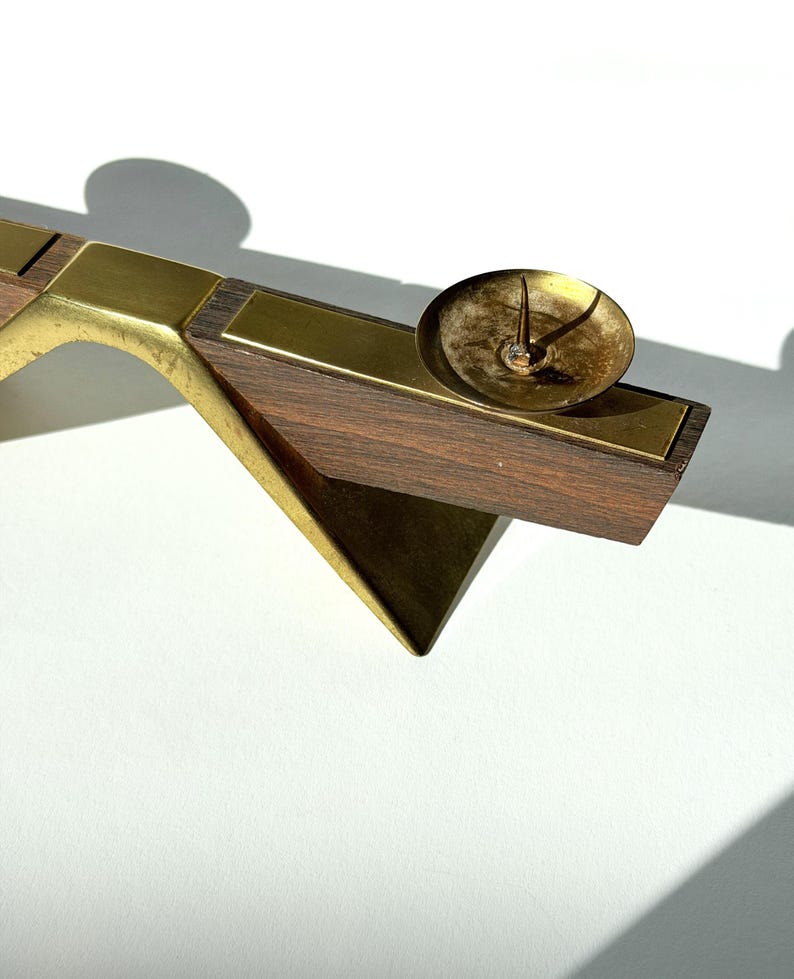 K&ouml;nnte beinhalten: Ein Kerzenhalter im Mid-Century-Modern-Stil mit Messing- und Holzdesign. Der Kerzenhalter verf&uuml;gt &uuml;ber eine kleine, runde Messingmulde f&uuml;r die Kerze und einen geometrischen Holzsockel. Das Holz ist dunkelbraun und das Messing hat einen warmen, goldenen Farbton.