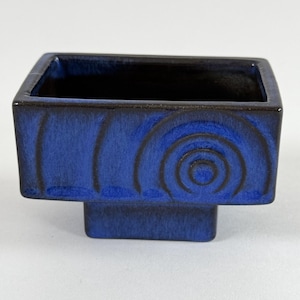 Könnte beinhalten: Ein blauer Keramik-Pflanztopf mit einem erhöhten Sockel und einem geometrischen Design. Der Topf hat eine dunkelblaue Glasur mit einem schwarzen Rand.