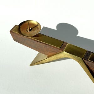 K&ouml;nnte beinhalten: Ein Vintage-Kerzenhalter mit Messing- und Holzdesign. Der Kerzenhalter hat eine kleine, runde Messing-Tasse, um eine Kerze zu halten, die auf einem rechteckigen Holzsockel steht. Der Sockel ist mit Messing akzentuiert und hat eine dreieckige St&uuml;tze.