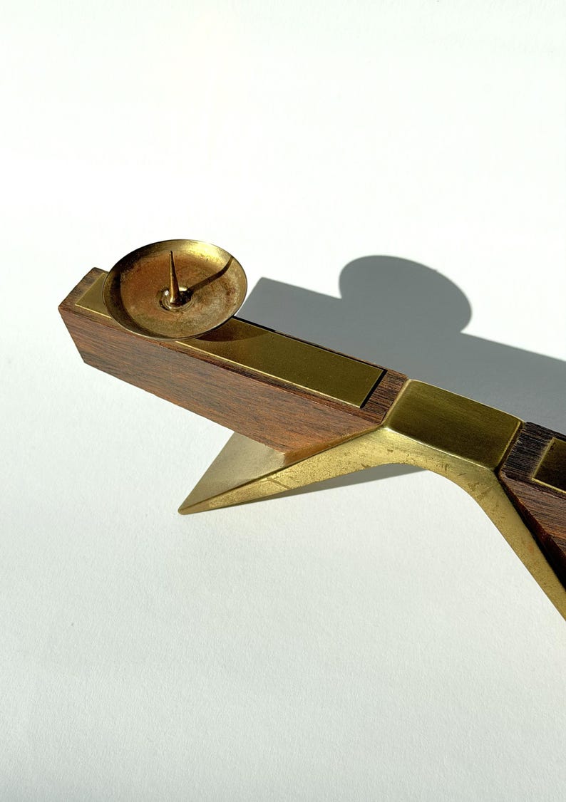 K&ouml;nnte beinhalten: Ein Vintage-Kerzenhalter mit Messing- und Holzdesign. Der Kerzenhalter hat eine kleine, runde Messing-Tasse, um eine Kerze zu halten, die auf einem rechteckigen Holzsockel steht. Der Sockel ist mit Messing akzentuiert und hat eine dreieckige St&uuml;tze.
