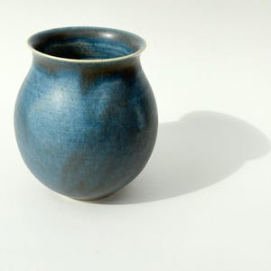 Pode incluir: Um vaso de cerâmica arredondado com um esmalte azul profundo, estrias mais escuras na cor carvão e uma silhueta curva e suave. O recipiente tem uma boca larga e aberta com uma borda subtil, exibindo um acabamento variado em tons de azul.