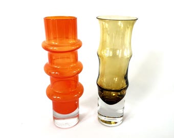 May include: Two glass vases. One is orange with a stacked design, the other is yellow-brown with a bamboo-like shape. Both have clear bases. The vases are set against a white background.