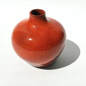 Op de afbeelding: Een ronde, oranjerode keramische vaas met een smalle hals. De vaas heeft een glanzende afwerking en een gespikkelde textuur. De binnenkant van de hals is donkerder rood. De vaas staat op een witte ondergrond.