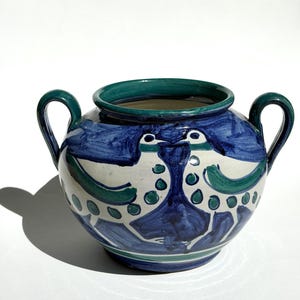 May include: A ceramic bowl with two handles, featuring a hand-painted design of two birds facing each other. The vessel is decorated with bold blue and green brushstrokes against a cream-colored background, with a solid dark green rim.
