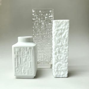 Könnte beinhalten: Drei dekorative Vasen. Die vordere Vase ist weiß mit einem geometrischen Muster. Die mittlere Vase ist klar mit einem strukturierten, rechteckigen Muster. Die dritte Vase ist weiß mit einem strukturierten, organischen Muster. Alle Vasen sind rechteckig.