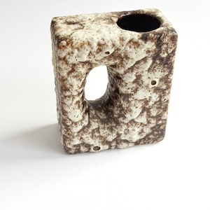 May include: A rectangular ceramic vase with a textured, cratered surface in mottled shades of cream and dark brown. The vase features a central oval cutout and a single circular opening at the top, with a dark interior finish.