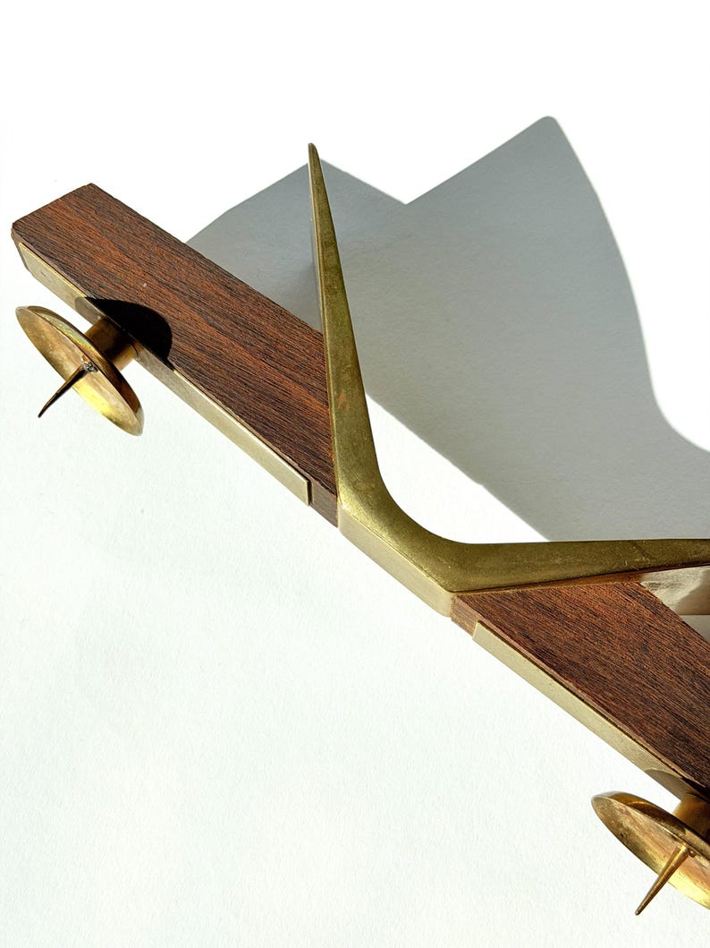 K&ouml;nnte beinhalten: Ein Vintage-Kerzenhalter mit Holzsockel und Messingdetails. Der Kerzenhalter hat zwei Messing-Kerzenschalen und ein hohes, gebogenes Messingelement. Das Holz ist dunkelbraun und das Messing hat einen warmen, goldenen Farbton.