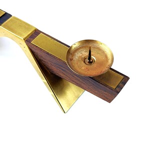 K&ouml;nnte beinhalten: Ein Vintage-Kerzenhalter mit einer Messing-Tasse und einem Dorn, der auf einem dunklen Holzsockel mit Messingakzenten steht. Der Halter hat ein geometrisches Design mit einem dreieckigen Sockel und einem rechteckigen Arm.