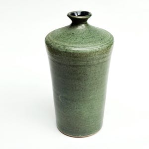 May include: A tall, cylindrical ceramic vase with a mottled sage green glaze. The vessel features a narrow, flared neck opening that reveals a dark, deep blue interior glaze. The exterior surface has a subtle, textured finish with horizontal lines.