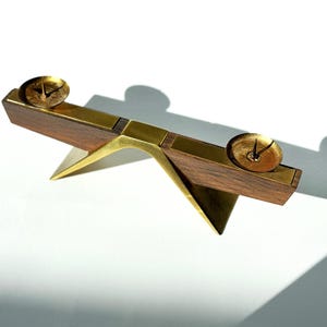 K&ouml;nnte beinhalten: Ein Kerzenhalter aus Holz und Messing mit zwei kreisf&ouml;rmigen Kerzenschalen. Der Halter hat ein Schaukelpferddesign mit einem dreieckigen Sockel und Messingakzenten. Das Holz ist dunkelbraun und die Kerzenschalen sind goldfarben.