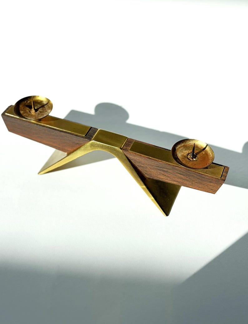 K&ouml;nnte beinhalten: Ein Kerzenhalter aus Holz und Messing mit zwei kreisf&ouml;rmigen Kerzenschalen. Der Halter hat ein Schaukelpferddesign mit einem dreieckigen Sockel und Messingakzenten. Das Holz ist dunkelbraun und die Kerzenschalen sind goldfarben.