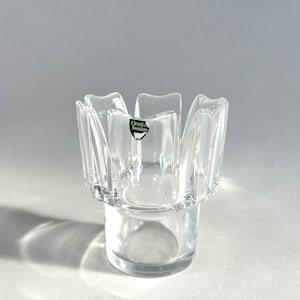 May include: Clear glass votive candle holder with a unique, flower-like design. The holder features a round base and a top that flares out with six petal-shaped sections. The Orrefors Sweden label is visible.