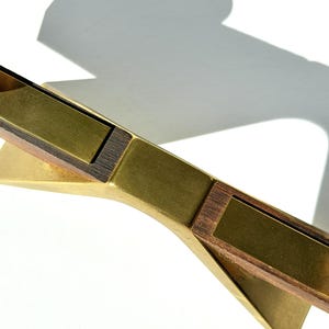 K&ouml;nnte beinhalten: Ein dekoratives Objekt aus Messing und Holz mit geometrischem Design. Das Objekt hat eine goldene Oberfl&auml;che mit rechteckigen Holzeinlagen. Der Schatten des Objekts wird auf eine wei&szlig;e Oberfl&auml;che geworfen.