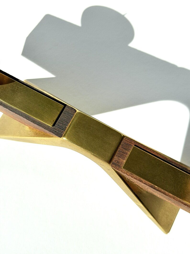 K&ouml;nnte beinhalten: Ein dekoratives Objekt aus Messing und Holz mit geometrischem Design. Das Objekt hat eine goldene Oberfl&auml;che mit rechteckigen Holzeinlagen. Der Schatten des Objekts wird auf eine wei&szlig;e Oberfl&auml;che geworfen.