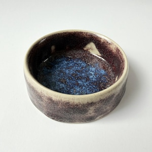 Könnte beinhalten: Eine kleine, runde Keramikschale mit strukturierter Oberfläche. Die Schale hat eine dunkelviolette Außenseite und eine blaue und braune Innenseite. Der Rand ist hellbeige. Diese handgefertigte Schale eignet sich zum Servieren kleiner Portionen.