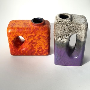 Puede incluir: Dos jarrones de cerámica. Uno es naranja y rojo con una superficie texturizada y una abertura circular. El otro es gris, morado y blanco con un diseño similar. Ambos tienen forma rectangular con un agujero.