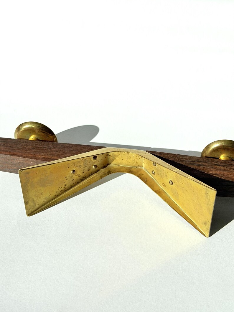 K&ouml;nnte beinhalten: Ein Eckwinkel aus Messing in V-Form, platziert &uuml;ber einem dunklen Holzelement mit zwei Messingkn&ouml;pfen. Die Halterung weist Schraubenl&ouml;cher und ein leicht gealtertes Aussehen auf, m&ouml;glicherweise Vintage. Der Hintergrund ist schlicht wei&szlig;.