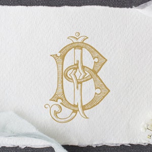 May include: A gold monogram with the letters B and C on a white paper. The paper is surrounded by a light blue ribbon, a pair of black scissors, a spool of ribbon, and small white flowers. The background is a dark gray surface.