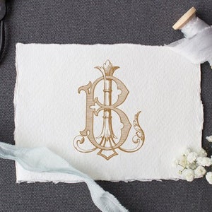 Può includere: Un monogramma dorato con la lettera "B" in un carattere decorativo. Il monogramma è su un pezzo di carta bianco con una texture ruvida. La carta è circondata da fiori bianchi, un rotolo di nastro bianco e un paio di forbici nere.