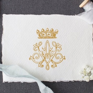 Op de afbeelding: Een gouden monogram met een kroon erboven, op een witte papieren achtergrond. Het monogram is omgeven door decoratieve versieringen. Het papier ligt op een grijs oppervlak.