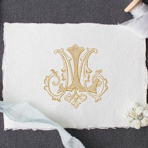 May include: A gold monogram design on a white paper with torn edges, accompanied by black scissors, a spool of gray ribbon, and small white flowers. A light blue ribbon and a smaller monogram are also present, all set against a dark gray background.