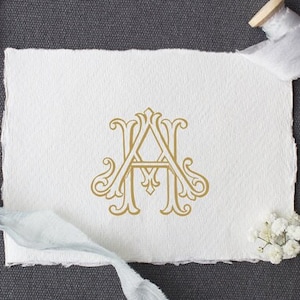 May include: A gold monogram featuring the letters "A" and "H" intertwined in a decorative script font. The monogram is displayed on a white piece of paper with a light blue ribbon and a spool of ribbon.
