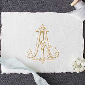 May include: A gold monogram with the letters "A M" in a decorative script font on a white background. The monogram is surrounded by a decorative border.