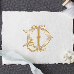 May include: A gold monogram with the letters "E" and "D" intertwined in a decorative script font. The monogram is on a white piece of paper with a rough texture.
