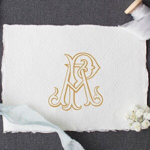May include: A white card with a gold monogram of the letters "A" and "R" intertwined. The card is on a grey surface with a spool of white ribbon and a sprig of white flowers.