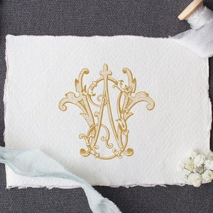 May include: A gold monogram with the letters "W" and "A" intertwined in a decorative, ornate design. The monogram is on a white piece of paper with a light blue ribbon and white flowers.