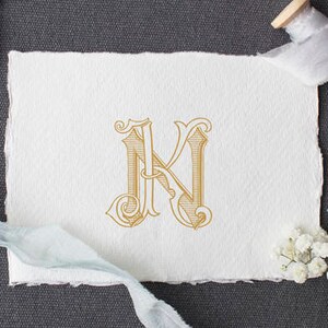 May include: A white card with a gold monogram of the letter "N" in a decorative script font. The card is surrounded by white ribbon, a spool of ribbon, and white flowers.
