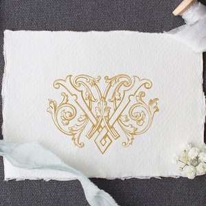 May include: A white card with a gold ornate monogram of the letters "W" and "W" intertwined. The card is on a gray surface with a pair of scissors, a spool of ribbon, and a small bouquet of white flowers.