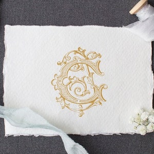May include: A gold-colored, ornate, decorative letter C monogram on a white paper background. The monogram is surrounded by a decorative floral design.