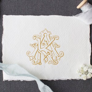 May include: A gold monogram with the letters "K" and "K" intertwined in a decorative, ornate design. The monogram is on a white piece of paper with a rough texture. The paper is on a gray surface.