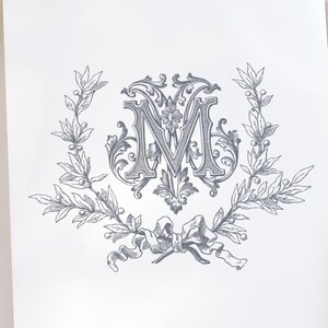 May include: A white card with a grey monogram featuring the letter "M" surrounded by a wreath of leaves and a bow.