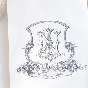 Puede incluir: Una invitación de boda blanca con un escudo gris y blanco que presenta las letras "N" y "J" rodeadas de diseños florales. El texto "Nancy & John" está impreso debajo del escudo.