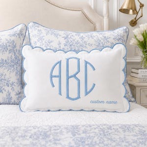 May include: A rectangular white throw pillow with a scalloped edge trimmed in light blue piping. The centre features a large, embroidered light blue monogram with the letters ABC, and the text custom name is embroidered in the bottom right corner.