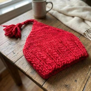 May include: A bright red knitted hat with a tassel, placed on a wooden table. The hat is made from thick, chunky yarn, and the tassel is the same red colour. A mug and a cream blanket are in the background.