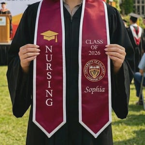 May include: A graduate in a black gown smiles, holding a maroon and white sash. The sash displays "NURSING" and "CLASS OF 2026" with a gold mortarboard graphic. The University of Calgary logo and the name "Sophia" are also visible.