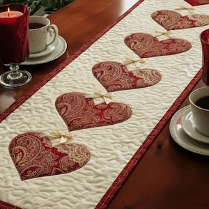May include: A cream quilted table runner with red paisley heart appliqués and a red border. Two red candles in glass holders and two cups of tea are on the wooden table. The runner is approximately 30 cm wide.