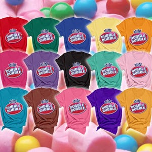 May include: A collection of fifteen short-sleeved t-shirts in various solid colors, each featuring the Dubble Bubble logo with a crown icon and the text America's Original Dubble Bubble printed in a red oval on the center of the chest.