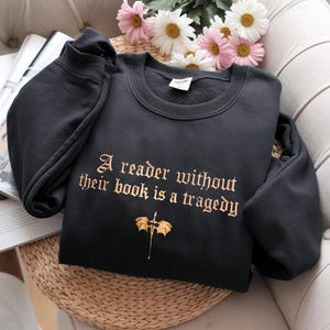 Puede incluir: Sudadera negra de cuello redondo con la cita "A reader without their book is a tragedy" en letras doradas. Una ilustración dorada de un dragón y una espada está debajo del texto. La sudadera está doblada sobre una superficie de mimbre.