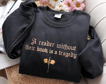 Può includere: Felpa nera girocollo con la citazione "A reader without their book is a tragedy" in lettere dorate. Un'illustrazione dorata di un drago e una spada è sotto il testo. La felpa è piegata su una superficie di vimini.