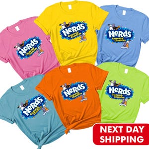 May include: Five short-sleeved crew neck t-shirts in pink, yellow, light blue, orange, and lime green. Each shirt features a central graphic with the text "NERDS Gummy Clusters" and "spooky" surrounded by colorful candy-themed illustrations.