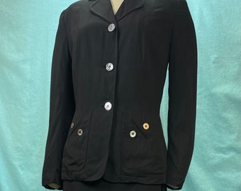 May include: A black skirt suit with a fitted jacket and a knee-length skirt. The jacket has a notched collar, four buttons down the front, and two flap pockets on the front. The skirt has a straight silhouette and a center back zipper.