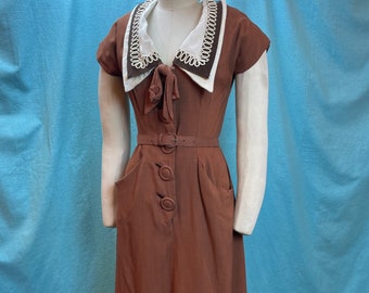Könnte beinhalten: Ein braunes Kleid mit einem weißen Kragen und braunen Verzierungen. Das Kleid hat einen Gürtel und Knöpfe an der Vorderseite.