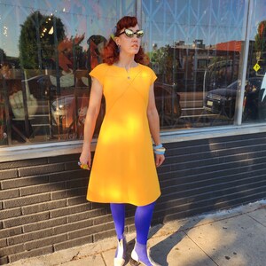 May include: A woman wearing a yellow dress, blue tights, and white shoes. She is standing in front of a shop with a window display.